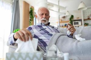 krank Alten Mann Überprüfung seine Temperatur Leiden von saisonal Grippe oder kalt Lügen auf Sofa lachend Leiden von saisonal Grippe oder kalt. . krank Senior Gefühl ungesund mit Grippe beim Zuhause foto