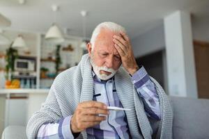 krank Alten Mann Überprüfung seine Temperatur Leiden von saisonal Grippe oder kalt Lügen auf Sofa lachend Leiden von saisonal Grippe oder kalt. . krank Senior Gefühl ungesund mit Grippe beim Zuhause foto