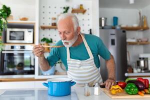 glücklich im Ruhestand Senior Mann Kochen im Küche. Ruhestand, Hobby Menschen Konzept. Porträt von lächelnd Senior Mann halten Löffel zu Geschmack Essen foto