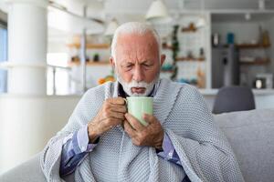 Senior Mann Leiden von Grippe Trinken Tee während Sitzung eingewickelt im ein Decke auf das Sofa beim heim. krank Mann mit ein kalt Lügen auf das Sofa halten ein Becher von heiß Tee. foto