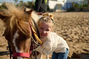 bezaubernd Kind Mädchen sanft umarmen ihr Pony Pferd, Stehen auf das sandig Strand foto