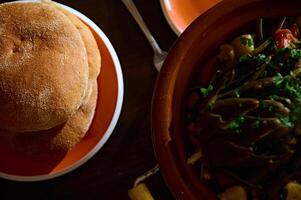 oben Aussicht von frisch gebacken Vollkorn Brot auf Keramik Platte. und Tajine mit gedämpft Gemüse auf ein hölzern Tisch. Essen Blog foto