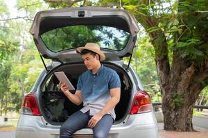 asiatisch Mann Sitzung zurück von Auto und halten Tabellen prüfen Ort und finden Platz zu Reise. glücklich Straße Ausflug und Ferien im Sommer- im das Landschaft. Konzept von Reise auf Freizeit, Straße Ausflug Ferien foto