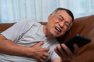 Senior männlich asiatisch drückt Hand zu seine Truhe hat ein Herz Attacke leidet von unerträglich Schmerzen im das Leben Zimmer, einsam Männer Versuchen mit das Smartphone zu Anruf zum Notfall Hilfe. foto