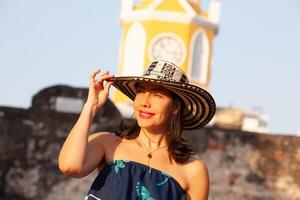 schön Frau tragen das traditionell kolumbianisch Hut namens Sombrero vueltiao beim das Uhr Turm auf das historisch Straßen von das Cartagena de Indien ummauert Stadt foto