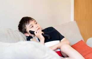 Kind Junge halten Fernbedienung Steuerung Sitzung auf Sofa, nah oben offen Porträt süß Schule Aufpassen Fernseher auf Wochenende.positiv Kind entspannend beim Zuhause nach Schule foto