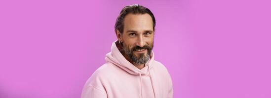 charmant verlockend gut aussehend bärtig stilvoll Erwachsene männlich Modell- Ohrring Rosa Kapuzenpullover lächelnd Erfreut ausdrücken Vertrauen Positivität Gefühl Glücklich amüsiert, Stehen lila Hintergrund reden beiläufig foto