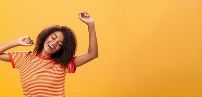 hüfthoch Schuss von entspannt und erleichtert glücklich afrikanisch amerikanisch jung weiblich Schüler im gestreift T-Shirt Dehnen Waffen oben Schließen Augen von Freude und lächelnd haben großartig Nickerchen Über Orange Hintergrund foto