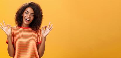 hüfthoch Schuss von Ausruhen abgehend Ruhe Freundin mit zuversichtlich aussehen im modisch gestreift T-Shirt zeigen okay oder Ausgezeichnet Geste und lächelnd breit versichern alles in Ordung Über Orange Hintergrund foto