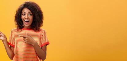 Porträt von erstaunt aufgeregt charismatisch dunkelhäutig jung ziemlich Mädchen mit afro Frisur im modisch gestreift T-Shirt zeigen links Erfreut und fasziniert posieren gegen Orange Hintergrund foto