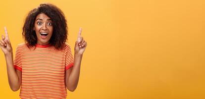 erstaunt glücklich begeistert süß afrikanisch amerikanisch Frau mit lockig Frisur im gestreift T-Shirt erziehen Index Finger zeigen oben Blick fasziniert und Erfreut beim Kamera Über Orange Mauer foto