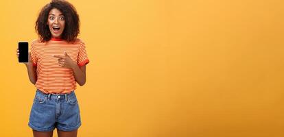 beeindruckt fasziniert stilvoll schlank afrikanisch amerikanisch lockiges Haar Frau im gestreift T-Shirt fallen lassen Kiefer von Staunen halten genial Smartphone zeigen beim Gerät Bildschirm Über Orange Mauer foto