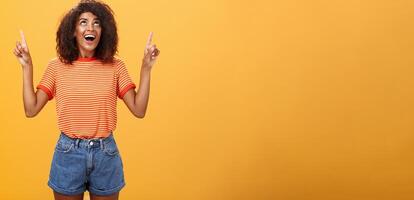 fasziniert beeindruckt und amüsiert gut aussehend charmant Afroamerikaner Frau mit afro Frisur im modisch T-Shirt und Denim kurze Hose zeigen und suchen oben mit interessiert aussehen Über Orange Mauer foto