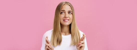 Frau haben Traum wollen Verlangen Kommen Sie wahr Kreuzung Finger zum gut Glück, Herstellung Wunsch beißen niedriger Lippe während Tagträumen Über Sache sie will Kaufen suchen beiseite posieren Über Rosa Hintergrund foto