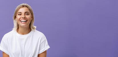 Nahansicht Schuss von froh charmant blond weiblich mit Erfreut und zufrieden Lächeln Stehen im Weiß T-Shirt Über lila Hintergrund Ausgaben Zeit im genial lustig Unternehmen foto