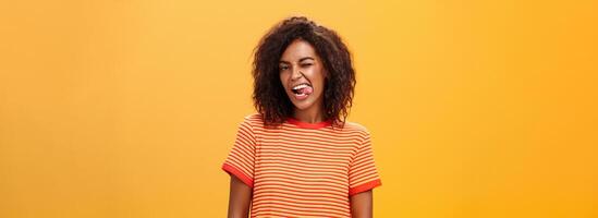 Porträt von gewagt und emotional zuversichtlich flirty Frau mit afro Frisur zwinkert freudig zeigen Zunge posieren sorglos und begeistert gegen Orange Hintergrund flirten mit heiß Kerl foto