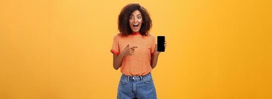 beeindruckt fasziniert stilvoll schlank afrikanisch amerikanisch lockiges Haar Frau im gestreift T-Shirt fallen lassen Kiefer von Staunen halten genial Smartphone zeigen beim Gerät Bildschirm Über Orange Mauer foto