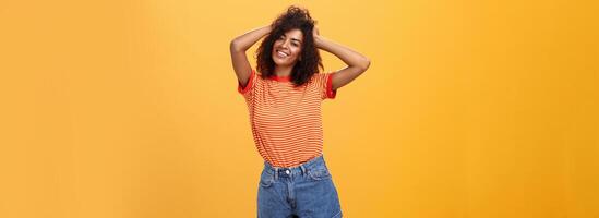 Mädchen Gefühl genial Herstellung cool Frisur posieren in der Nähe von Spiegel Gefühl froh zu haben Datum heute Abend halten Hände auf lockig Haar lächelnd freudig und zärtlich Stehen gegen Orange Hintergrund foto