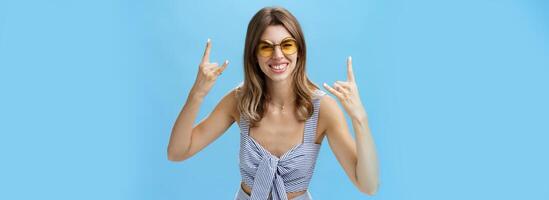 Mädchen bereit zu Rock 'n' Roll diese Party tragen Beste passend Outfit von Kleiderschrank und cool Sonnenbrille zeigen Felsen Gesten, lächelnd aufgeregt haben Spaß Hören Musik- auf Konzert Über Blau Mauer foto