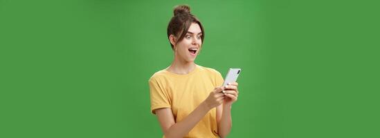 Frau lesen überraschend befriedigend Botschaft im Smartphone Öffnung Mund von Aufregung, lächelnd erstaunt suchen erstaunt beim Handy Bildschirm posieren gegen Grün Hintergrund im beiläufig Gelb T-Shirt foto