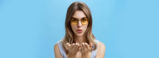 Nahansicht Schuss von feminin und zärtlich charmant Frau im modisch Gelb Sonnenbrille falten Lippen halten Palmen in der Nähe von Mund zu senden Luft Kuss beim Kamera Blick sinnlich nach vorne Über Blau Hintergrund foto