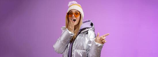 erstaunt sprachlos überrascht attraktiv stilvoll blond Freundin im Sonnenbrille, Silber glänzend Jacke Hut fallen Kiefer beeindruckt erweitern Augen schockiert zeigen links erstaunt, lila Hintergrund foto