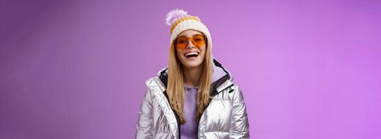 stilvoll freundlich charismatisch blond Frau im Silber glänzend Jacke Hut Sonnenbrille bereit lernen Snowboarden lächelnd Lachen glücklich haben Spaß schneebedeckt Erholung Erholungsort, Stehen lila Hintergrund foto