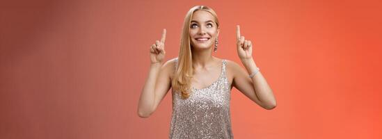 amüsiert Herrlich zärtlich elegant europäisch Frau blond lange Frisur im Silber glänzend Abend Kleid aussehen zeigen oben neugierig Blick Aufregung sehen gewünscht Ding, Stehen rot Hintergrund froh foto