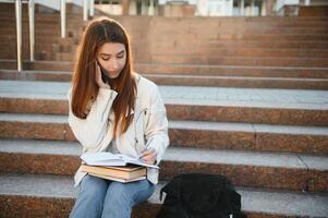 Porträt von erfolgreich Student. glücklich Teenager Mädchen posieren halten Bücher lächelnd suchen beim Kamera Stehen in der Nähe von modern Universität Gebäude außen. Studium, modern Bildung foto