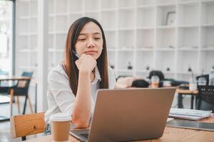 Geschäft Analytik Konzept, nachdenklich asiatisch Geschäftsfrau mit Stift im Hand Arbeiten auf ein Laptop, mit ein Kaffee Tasse und Unterlagen auf das Tabelle im ein Cafe. foto