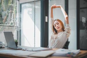 Geschäft Analytik Konzept, ein Frau im ein Licht Bluse nimmt ein Moment zu strecken und entspannen beim ihr Arbeit Schreibtisch, inmitten ein beschäftigt Büro Tag. foto