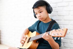 asiatisch Junge tragen Kopfhörer mit schwarz Musik- sitzen und abspielen Gitarre im das Haus. Musik- Lernen Konzept, Musik- Kompetenz Ausbildung. Kopieren Raum foto