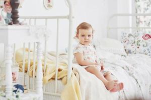 Babymädchen in süßem Kleid, das im Bett spielt und mit Spielzeug zu Hause spielt. weißes Vintage-Kinderzimmer. Kindheit Konzept foto