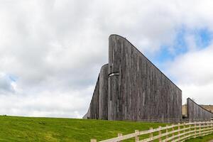 modern hölzern die Architektur mit gebogen Design gegen ein wolkig Himmel, umgeben durch Grün Gras und ein Zaun. foto