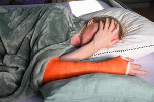 Frau mit ein gebrochen richtig Arm mit ein Orange Glasfaser Gips Besetzung schläft im Bett mit ein Kissen unter das Besetzung foto