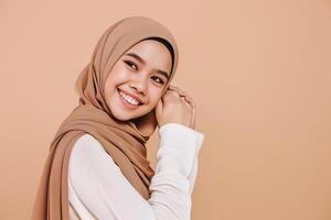 ai generiert schön asiatisch jung Muslim Frau tragen Kopftuch, lächelnd und suchen beim Kamera auf Beige Hintergrund foto