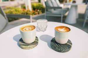 zwei Tassen von köstlich Kaffee und ein Glas von Eis Stand auf ein Weiß Tabelle im ein Sommer- Cafe auf ein sonnig Tag foto