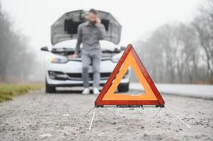 Mann verwenden ein Handy Anruf Garage im Vorderseite von das öffnen Kapuze von ein gebrochen Auto auf das Straße im das Wald. Auto Nervenzusammenbruch Konzept. foto