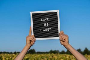 Hände von Demonstrant mit Text speichern das Planet auf schwarz Tafel auf Hintergrund von Sonnenblume Feld. Wiederverwendung reduzieren recyceln Konzept. protestieren zum Natur Klima Streik Freiwillige Protest gegen Erde Verschmutzung, global Erwärmen foto