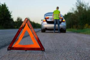 unkenntlich traurig Treiber im reflektierend Weste. männlich Treiber Stehen in der Nähe von gebrochen Auto mit öffnen oben Haube. rot Dreieck zu warnen andere Straße Benutzer von Auto Nervenzusammenbruch oder Motor Fehler halt beim Landschaft Autobahn. Notfall der Verkehr Situation foto