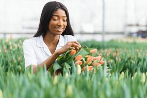 schön jung lächelnd afrikanisch amerikanisch Mädchen, Arbeiter mit Blumen im Gewächshaus. Konzept Arbeit im das Gewächshaus, Blumen. foto