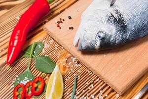 frisch roh Dorado Fisch. Meer Brachsen, Lügen auf ein hölzern Tafel auf ein Tabelle mit Kräuter und Salz- und Gewürze. Dorado und Zutaten zum Kochen auf das Tisch. Nahansicht foto