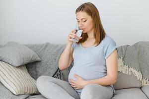 schwanger Frau mit Glas von Wasser Sitzung auf Sofa. jung erwartungsvoll blond sich ausruhen beim heim, Kopieren Raum. Schwangerschaft, Gesundheitspflege, Durst Konzept foto