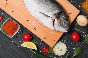 frisch roh Dorado Fisch auf Backen Papier mit Zitrone, Pfeffer, Tomaten und verschiedene Gewürze auf hölzern Hintergrund mit Kopieren Raum. foto