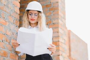 Konstruktion Ingenieur. Mädchen mit Konstruktion Dokumentation. ein Frau im ein Weiß schwer Hut gegen das Dach von ein Gebäude. Konstruktion von ein Neu Haus foto