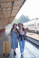 zwei jung asiatisch freunde Mädchen mit Rucksäcke beim Eisenbahn Bahnhof warten zum Zug, zwei schön Frauen Gehen entlang Plattform beim Zug Bahnhof foto