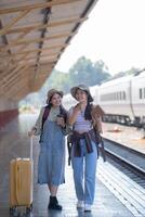 zwei jung asiatisch freunde Mädchen mit Rucksäcke beim Eisenbahn Bahnhof warten zum Zug, zwei schön Frauen Gehen entlang Plattform beim Zug Bahnhof foto