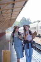 zwei jung asiatisch freunde Mädchen mit Rucksäcke beim Eisenbahn Bahnhof warten zum Zug, zwei schön Frauen Gehen entlang Plattform beim Zug Bahnhof foto