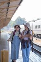 zwei jung asiatisch freunde Mädchen mit Rucksäcke beim Eisenbahn Bahnhof warten zum Zug, zwei schön Frauen Gehen entlang Plattform beim Zug Bahnhof foto