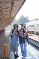 zwei jung asiatisch freunde Mädchen mit Rucksäcke beim Eisenbahn Bahnhof warten zum Zug, zwei schön Frauen Gehen entlang Plattform beim Zug Bahnhof foto